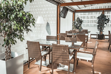 Attractive transparent glass terrace of the modern restaurant. Stylish interior the wicker tables and comfortable chairs in beige shades. Fashionable Mediterranean style.