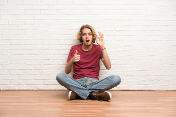 Fototapeta na wymiar Blonde man sitting on the floor showing ok sign and thumb up gesture