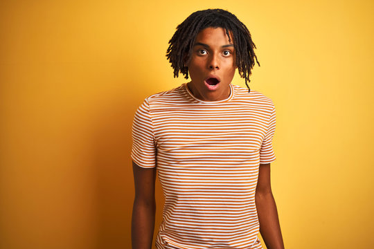 Afro man with dreadlocks wearing striped t-shirt standing over isolated yellow background afraid and shocked with surprise and amazed expression, fear and excited face.