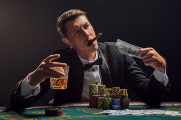 Handsome emotional man is playing poker sitting at the table in casino against a white spotlight.