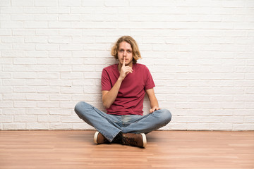 Blonde man sitting on the floor showing a sign of silence gesture putting finger in mouth