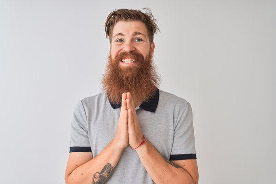 Young redhead irish man wearing grey polo standing over isolated white background praying with hands together asking for forgiveness smiling confident.
