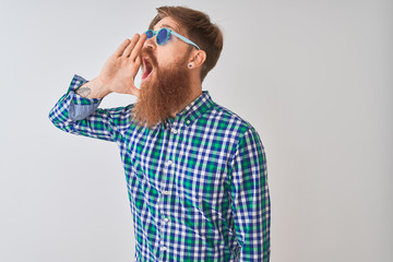 Young redhead irish man wearing casual shirt and sunglasses over isolated white background shouting and screaming loud to side with hand on mouth. Communication concept.