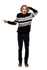 Full-length shot of Blonde man surprised and pointing finger to the side over isolated white background
