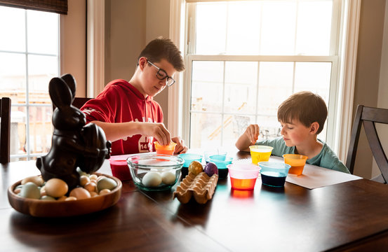 Two boys coloring Easter eggs with containers of dye at wooden table.
