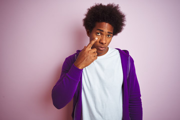 Young african american man wearing purple sweatshirt standing over isolated pink background Pointing to the eye watching you gesture, suspicious expression