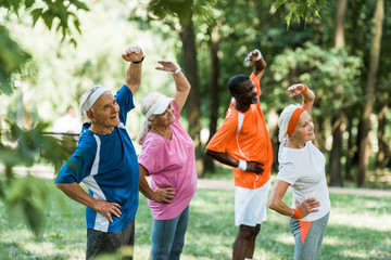 selective focus of cheerful multicultural retired men and women standing with hands on hips while...