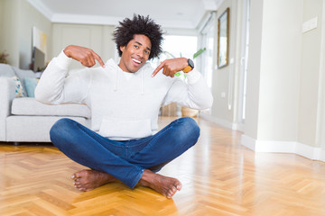 African American man sitting on the floor at home looking confident with smile on face, pointing...