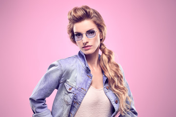 Fashion. Gorgeous long-haired blonde woman in Stylish denim jacket. Glamour Lady in Trendy Outfit, blue Sunglasses. Beautiful fashionable Model Girl with trendy hairstyle, makeup in Studio on Pink