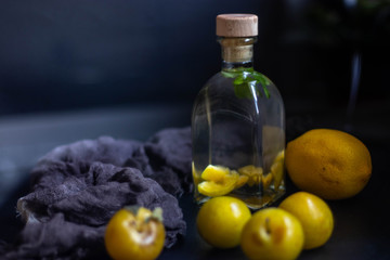 Plum tincture in a square bottle. Closed wooden stopper. Near yellow plums and a big lemon. All on a black background.