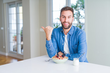 Handsome man eating cereals for breakfast at home smiling with happy face looking and pointing to the side with thumb up.