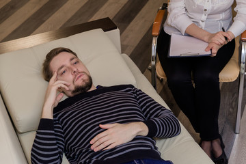 Fototapeta premium Young man at the psychotherapist, lying on couch, emotionally speaking, discussing with psychotherapist his problems, practitioner making some notes