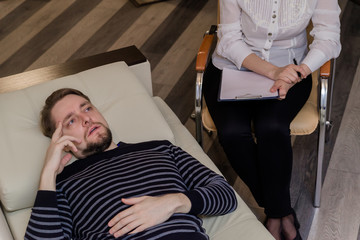 Young man at the psychotherapist, lying on couch, emotionally speaking, discussing with psychotherapist his problems, practitioner making some notes