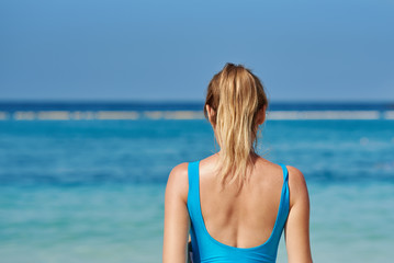 Fototapeta na wymiar Blonde girl in swimsuit against blue sea water on beach during summer vacation. Back view.