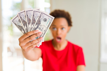 Young african american woman holding bank notes of fifty dollars scared in shock with a surprise face, afraid and excited with fear expression