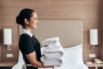 side view of smiling maid holding pile of folded towels near bed in hotel room - obrazy, fototapety, plakaty