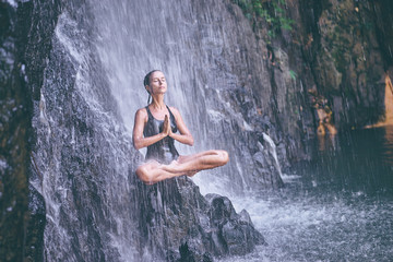 Wellness spa, vacation and yoga meditation concept. Pretty young woman in swimsuit sitting in lotus position on the rock under tropical waterfall.