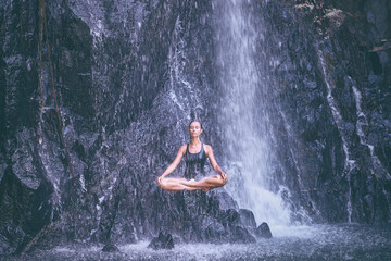 Wellness spa, vacation and yoga meditation concept. Pretty young woman in swimsuit sitting in lotus position on the rock under tropical waterfall.