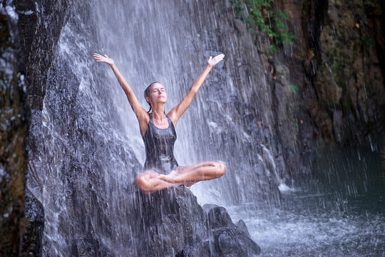 Young woman with open arms in lotus position under waterfall in tropical nature. Wellness spa and yoga meditation concept in holiday nature.
