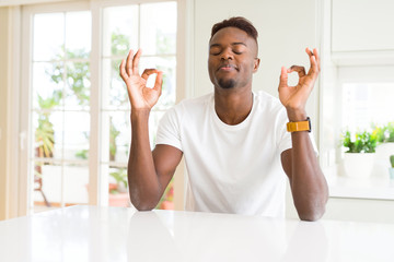Handsome african american man on white table at home relax and smiling with eyes closed doing meditation gesture with fingers. Yoga concept.