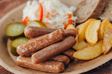 Tasty sausages with fried potatoes, cucumber, cabbage and chlobe in a paper plate on a wooden table.