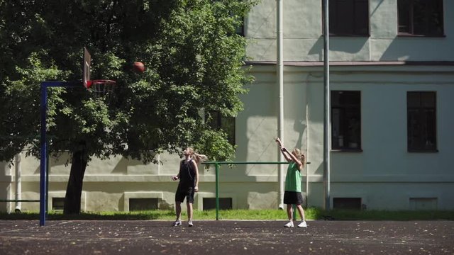 Side view of two female college friends playing basketball on street court to practice skills. Athletic girl making successful shot into hoop and scoring goal, wide slow motion shot