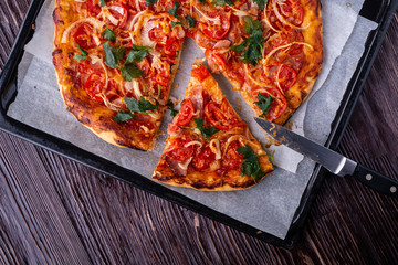 Freshly cooked hot pizza and slice with cheese ham meat tomato onion parsley homemade on parchment paper in baking sheet tray recipe with kitchen knife on dark wooden table