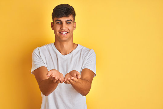 Young indian man wearing white t-shirt standing over isolated yellow background Smiling with hands palms together receiving or giving gesture. Hold and protection