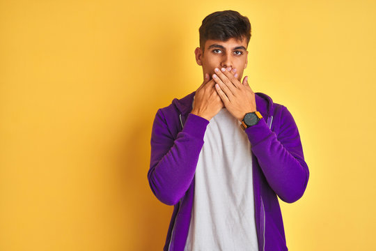 Young indian man wearing purple sweatshirt standing over isolated yellow background shocked covering mouth with hands for mistake. Secret concept.