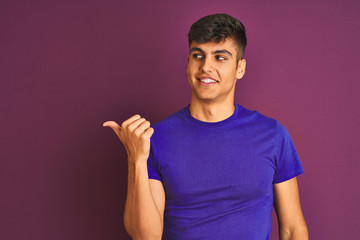 Young indian man wearing t-shirt standing over isolated purple background smiling with happy face looking and pointing to the side with thumb up.