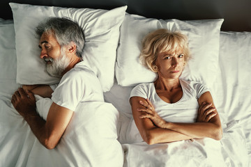 Overhead of senior couple in the bed. Pretty aged woman is angy with her sleeping husband.