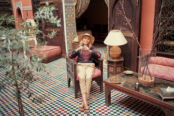 Traveling by Morocco. Happy young woman in hat relaxing in traditional riad interior in medina.