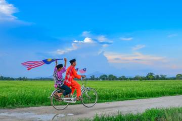 independence Day concept - Two happy young local boy riding old bicycle at paddy field holding a Malaysian flag