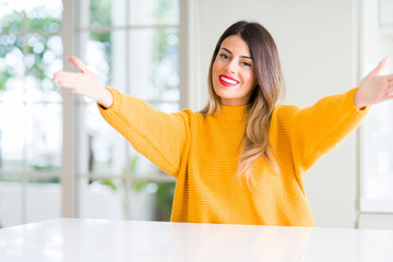 Young beautiful woman wearing winter sweater at home looking at the camera smiling with open arms for hug. Cheerful expression embracing happiness.