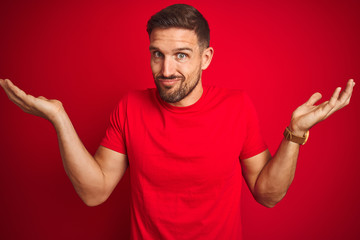 Young handsome man wearing casual t-shirt over red isolated background clueless and confused expression with arms and hands raised. Doubt concept.