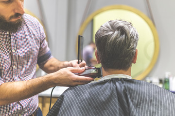 Barber styling hair of his client in front of mirror by using comb and clipper at barbershop.