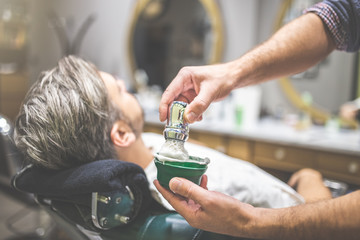 Close up of barber hands preparing foam by using shaving brush and bowl. Client in armchair in the background.