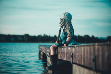 The child in the gas mask is sitting on the platform at the lake. Environment pollution.