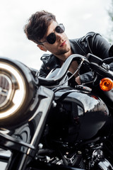selective focus of handsome motorcyclist in leather jacket looking away while sitting on motorcycle
