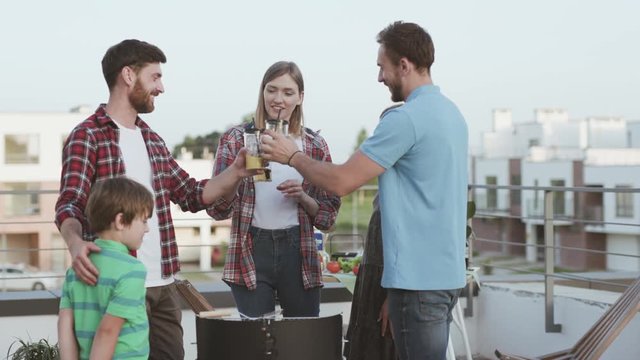 Delighted family with kids enjoying life hanging out together at a party. Young people clinking glasses cooking barbeque food on the roof of the building on sunny summer evening.