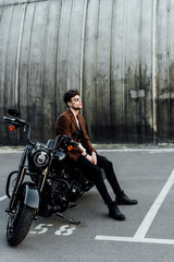 full length view of motorcyclist ion brown jacket sitting on motorcycle, resting and looking away