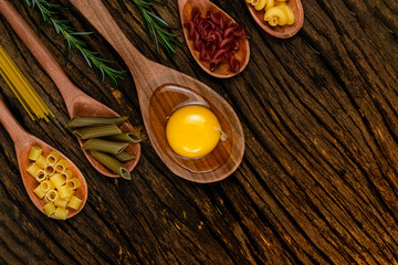 Different pasta types in wooden spoons on the table. Top view. Space for text. Old wooden background.