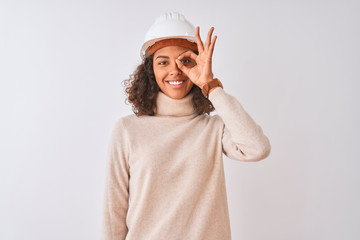 Young brazilian architect woman wearing security helmet over isolated white background doing ok gesture with hand smiling, eye looking through fingers with happy face.