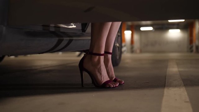 Close-up of slim female legs in high heeled shoes stepping out of own automobile parked in underground supermarket parking. Pretty woman in white dress and high heels getting out of car and walking