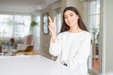 Young beautiful woman at home on white table smiling with happy face winking at the camera doing victory sign. Number two.