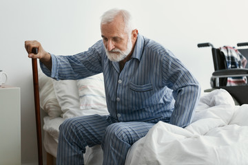 Sad senior man with Alzheimer's disease holding walking stick and sitting on his bed in nursing home