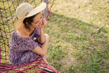 Young woman holding her summer hat while sitting in net hammock in her backyard in sunny day smiling happy