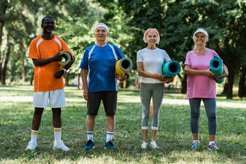 happy retired and multicultural pensioners holding fitness mats while standing on green grass in park