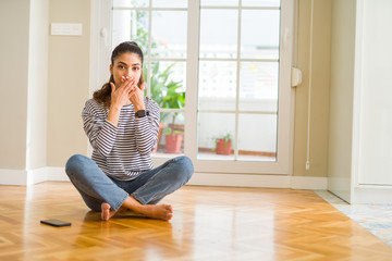Young beautiful woman sitting on the floor at home shocked covering mouth with hands for mistake. Secret concept.