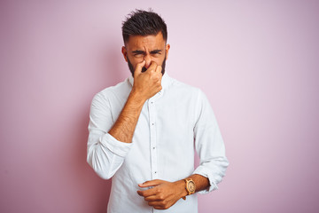 Young indian businessman wearing elegant shirt standing over isolated pink background smelling something stinky and disgusting, intolerable smell, holding breath with fingers on nose. Bad smells 